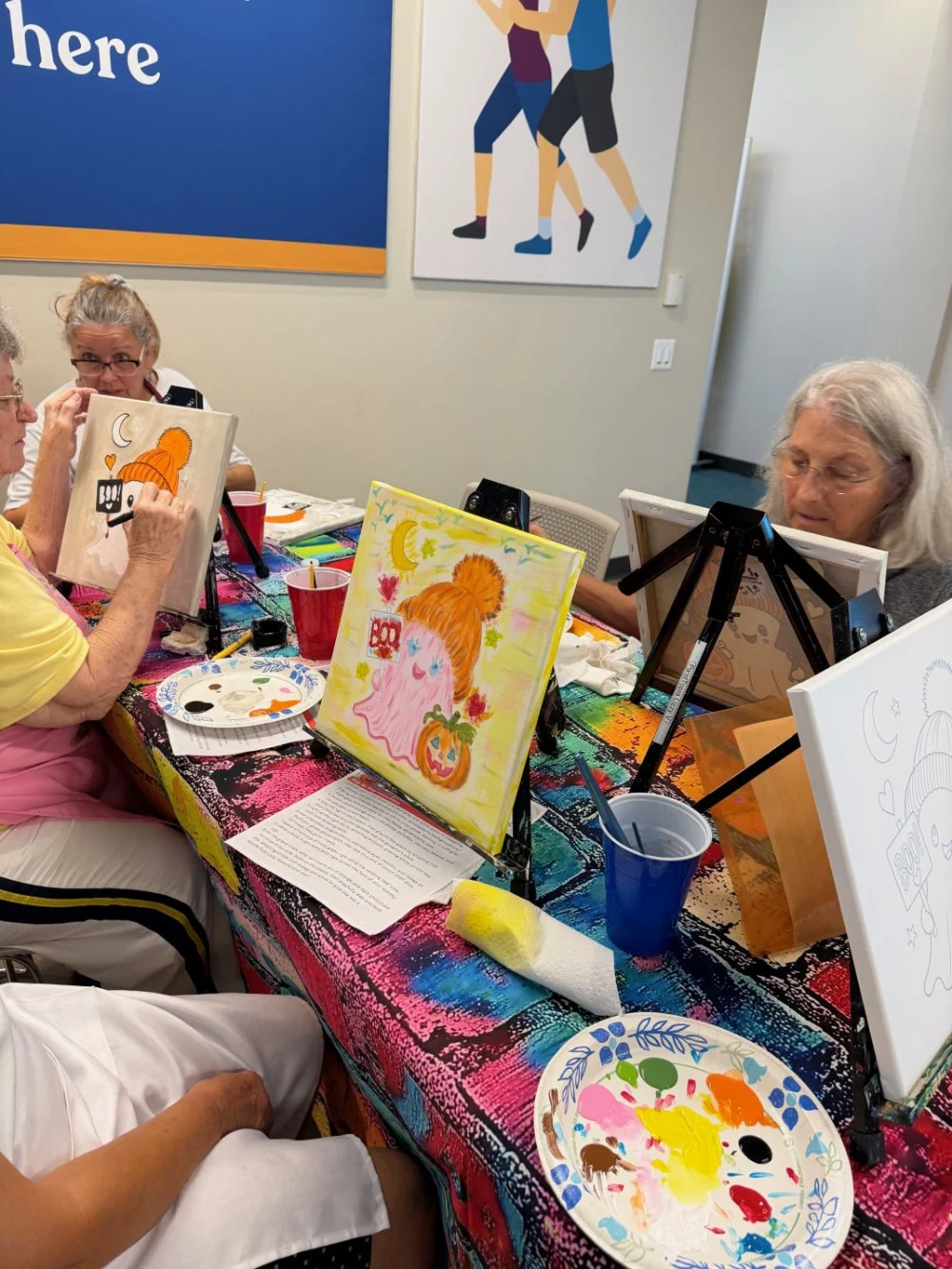 Group of women painting Halloween-themed artwork at a table in a room, with paintings of pumpkins and ghosts, and supplies like paint, brushes, and plates.