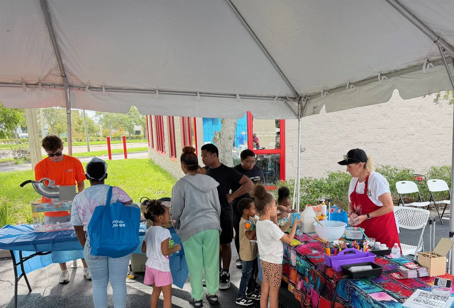Children and adults at an outdoor craft and activity booth under a white canopy tent, with a vendor in a red apron and a woman in a gray hoodie looking on.