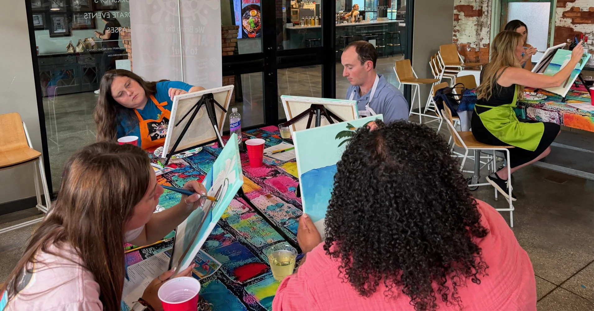 People painting at a group art event in a cafe or studio, with canvases on easels, colorful tablecloth, and drinks.