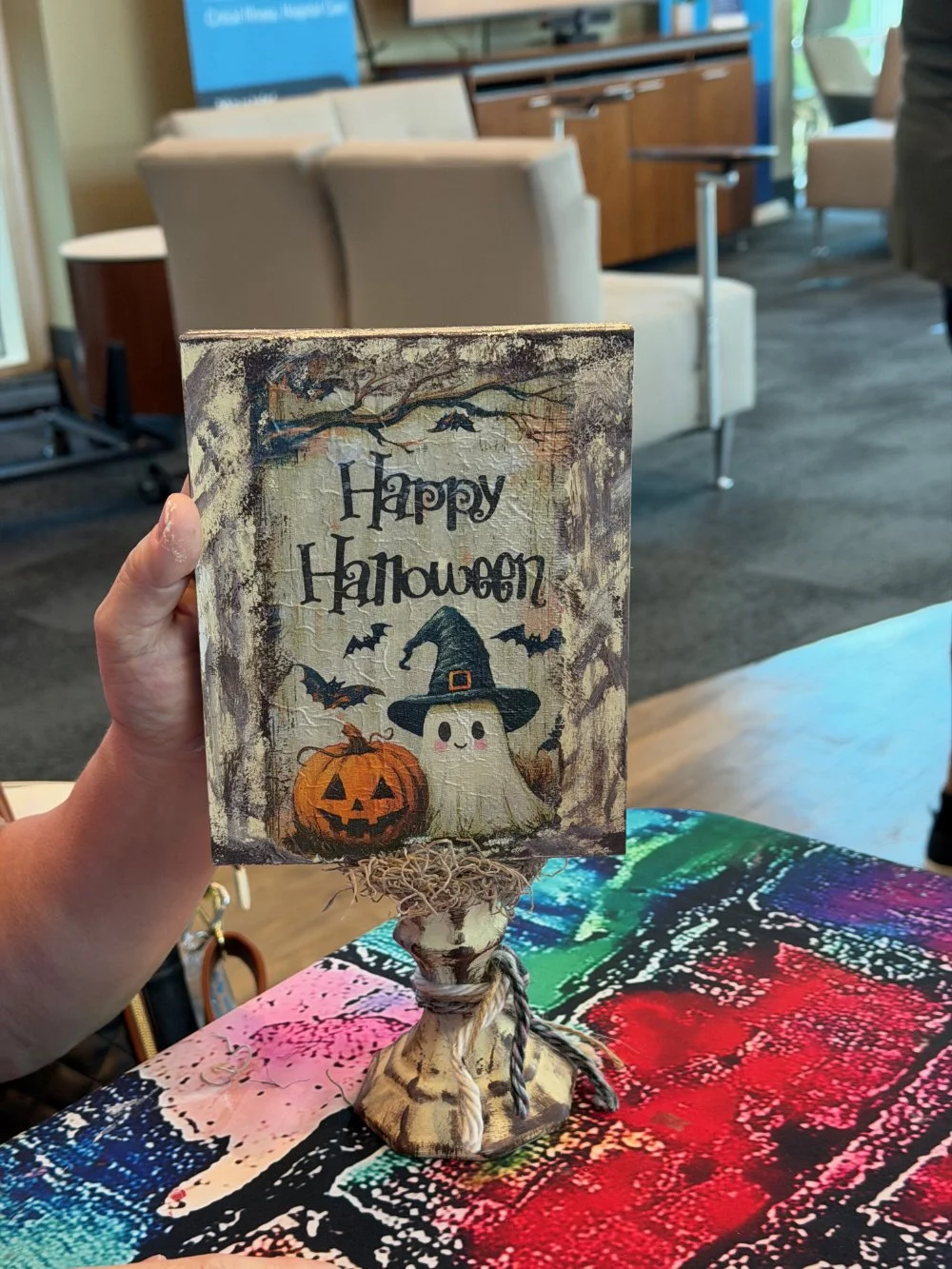 A Halloween-themed decorative sign with a ghost wearing a witch hat, a carved pumpkin, and bats, holding a sign that reads 'Happy Halloween' in a cozy indoor setting.
