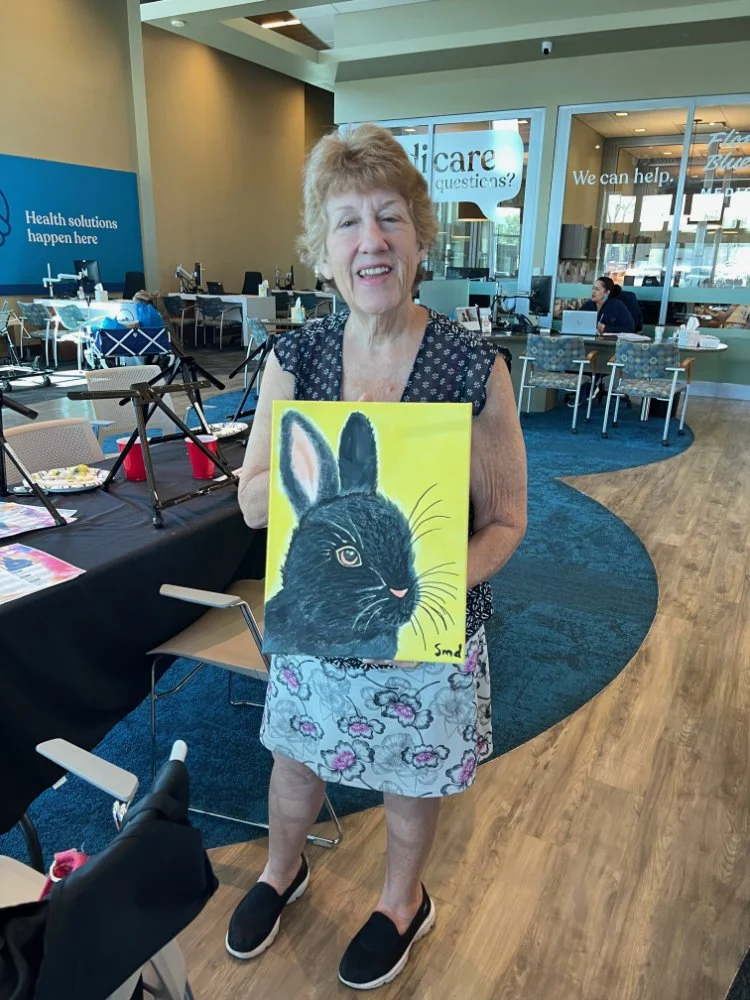An elderly woman holding a painted picture of a black rabbit with a yellow background in a cafe or community space.