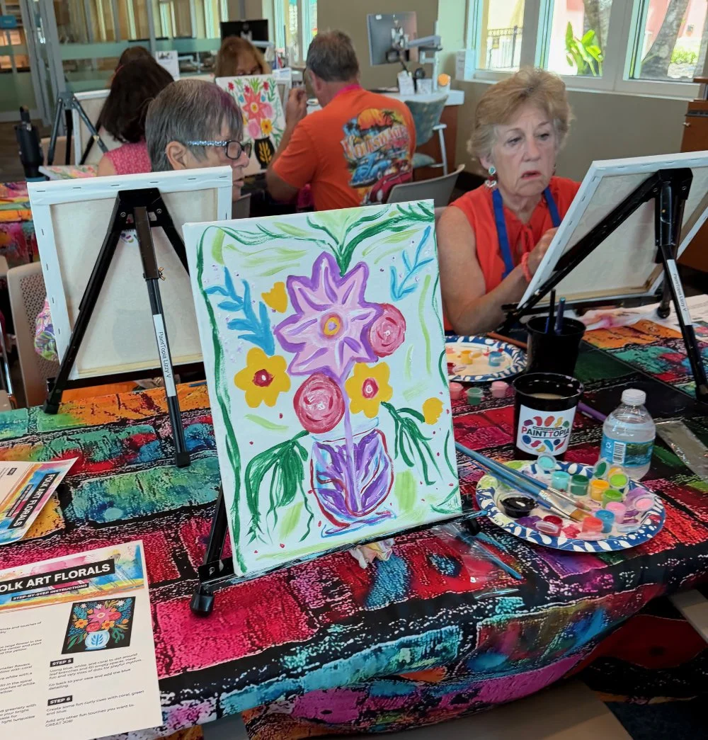 A group of people participating in a painting class, with an older woman in orange in the foreground painting on a canvas. The table is covered with a colorful, patterned cloth, and there is a finished floral painting in the center, along with paint 