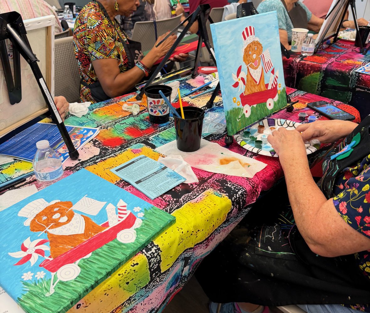 People painting colorful pictures of dogs dressed as Uncle Sam on canvases at a group art event, with painting supplies on the table.