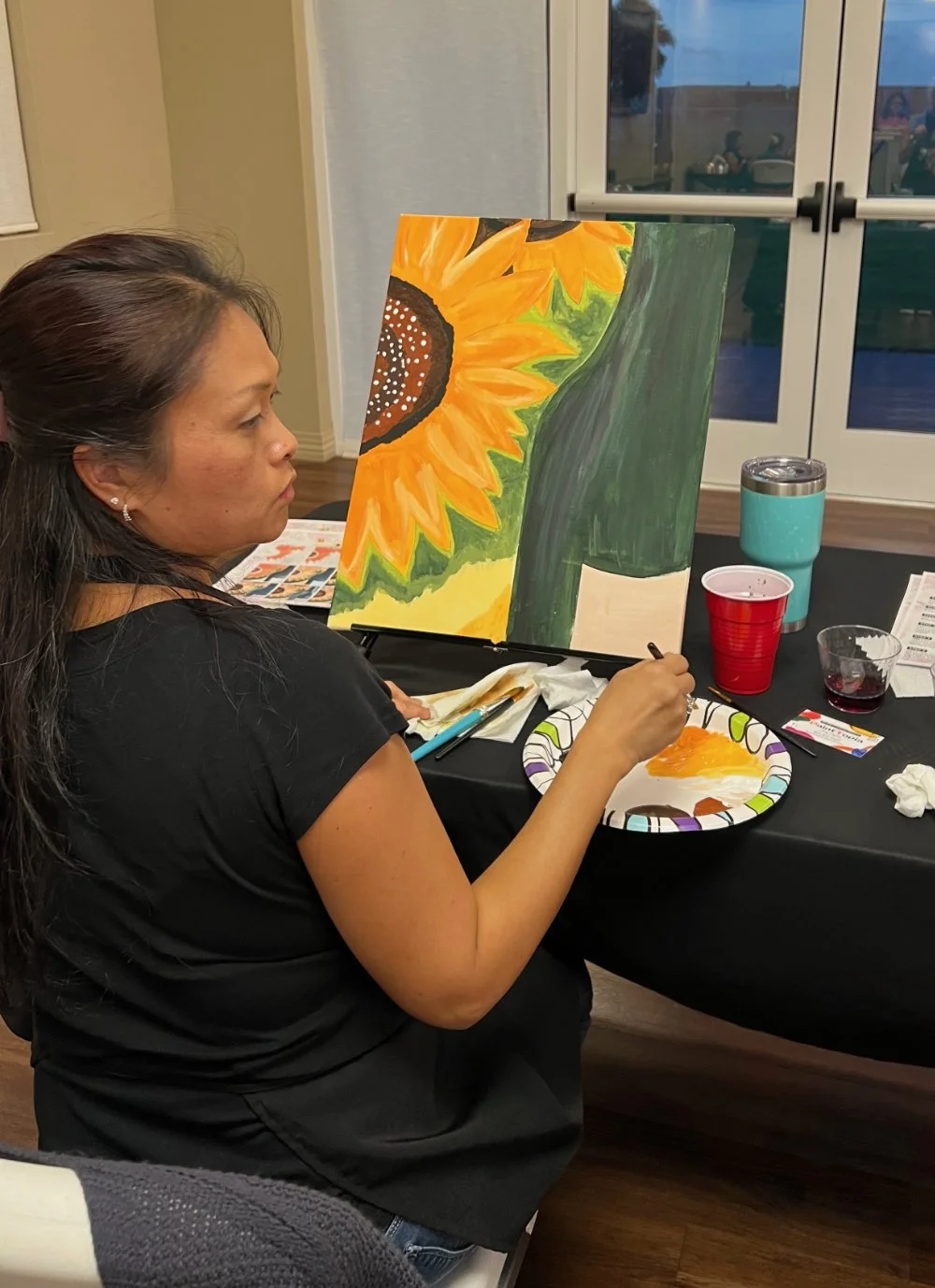 A woman painting a sunflower on a canvas at a table with art supplies, cups, and paint.