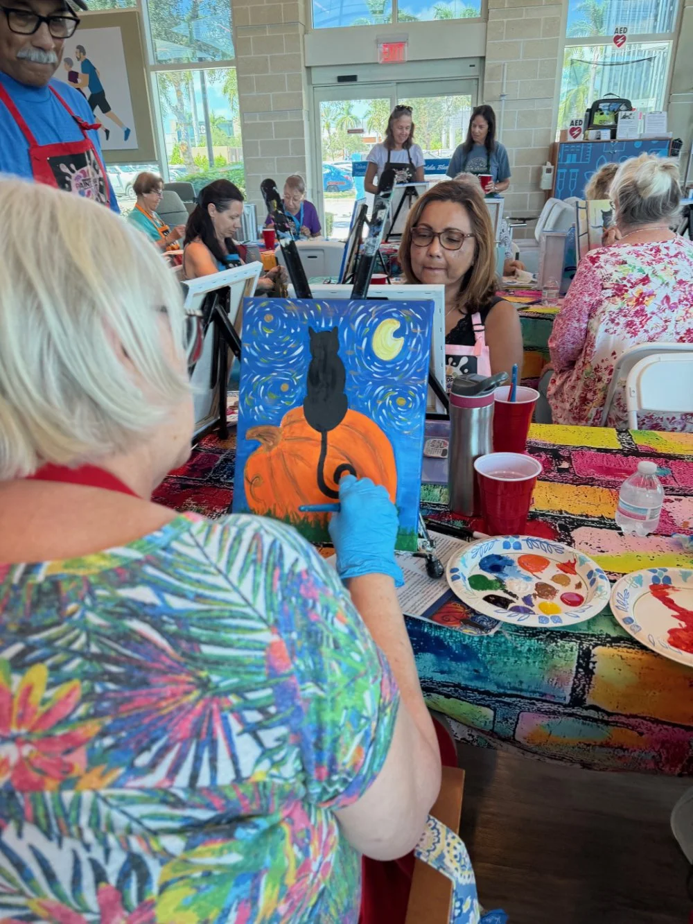 A group of people participating in a painting class, with one woman painting a picture of a black cat sitting on a pumpkin against a starry night sky. The setting is a bright, indoor space with large windows, and colorful art supplies and drinks are 