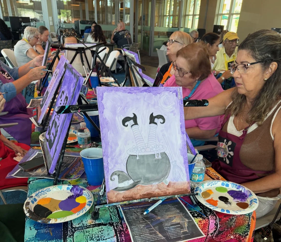Group of women participating in a painting class, working on individual canvases, with a completed painting of a bubbling cauldron with a wizard's hat nearby, in a bright indoor setting.