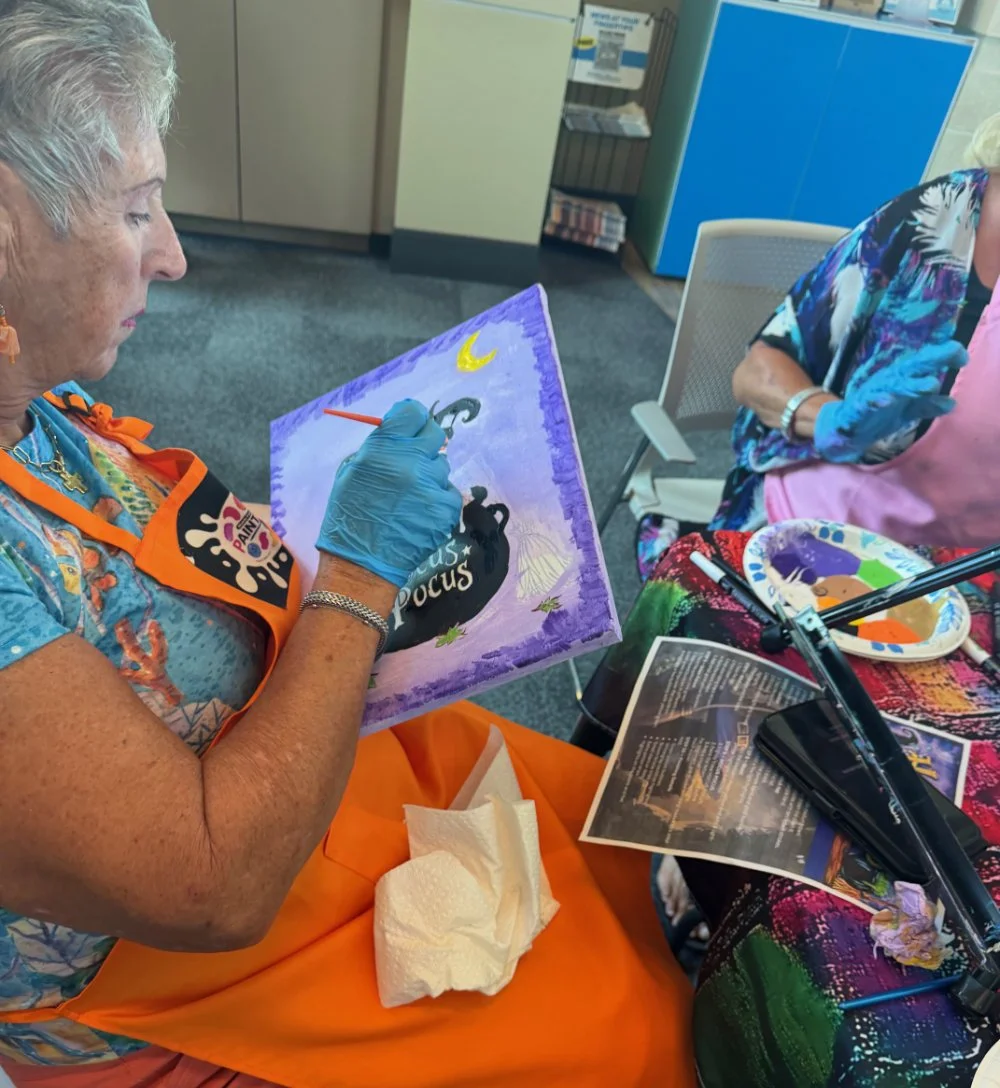 An elderly woman painting on a canvas at a crafting event, surrounded by art supplies and other participants.