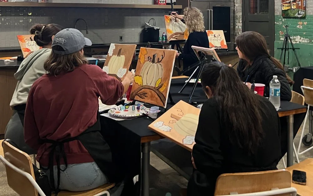 Group of people painting autumn-themed canvases with pumpkins and leaves at a craft class, in a cozy studio with an industrial look.