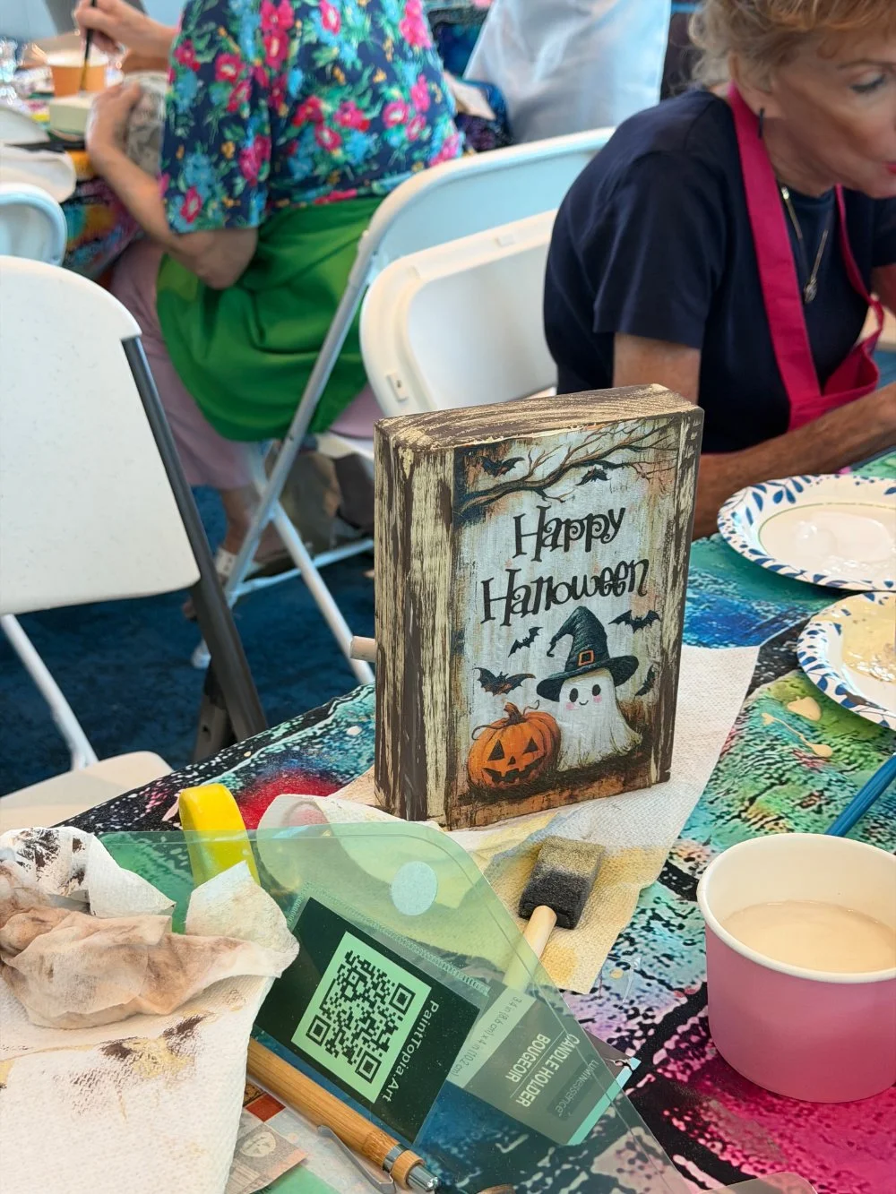Decorative Halloween sign featuring a ghost with a witch's hat, a pumpkin, and bats with the text 'Happy Halloween' on a colorful party table.