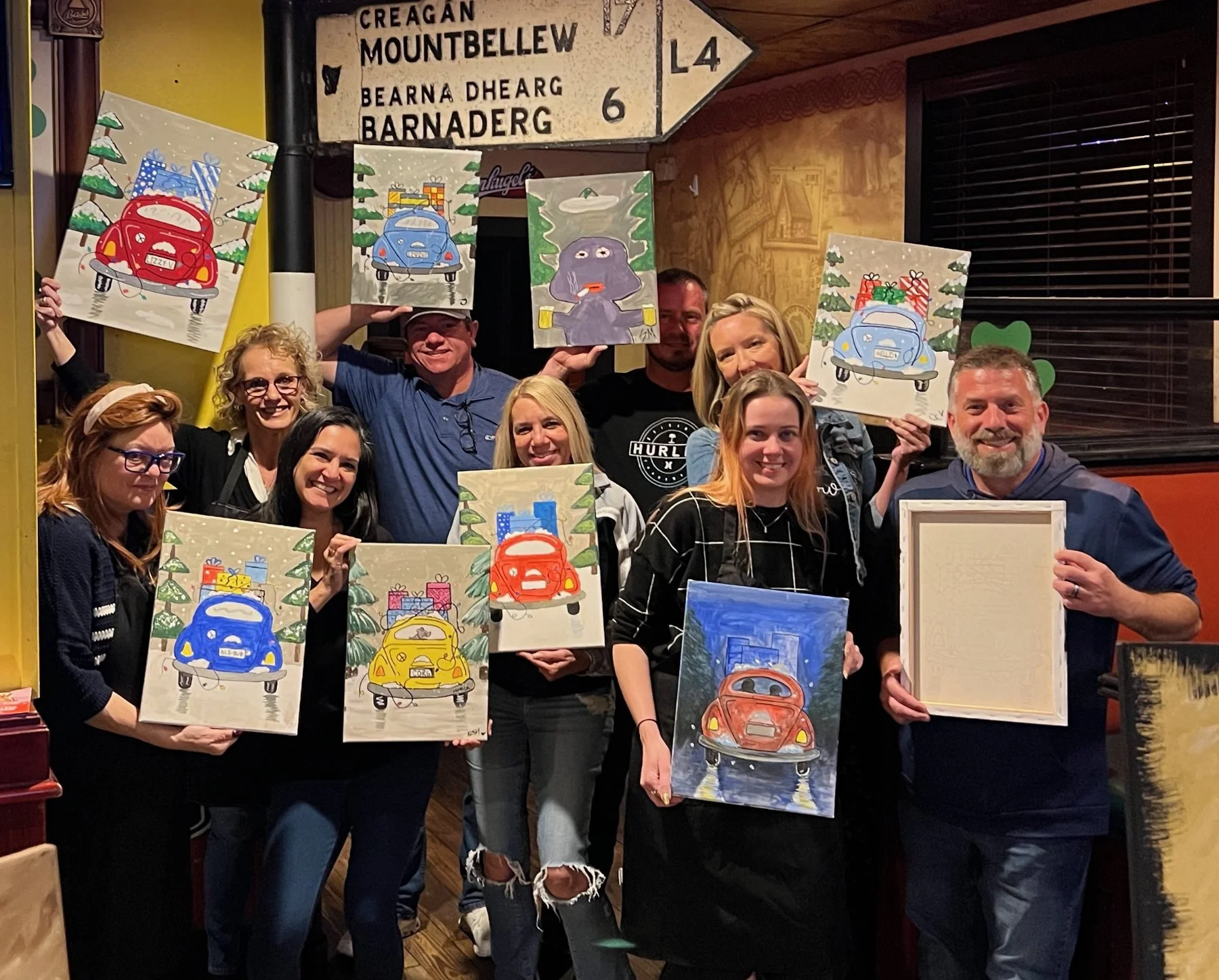 Group of people celebrating a painting event, holding up colorful artwork featuring cars and Christmas trees in a cozy indoor setting.