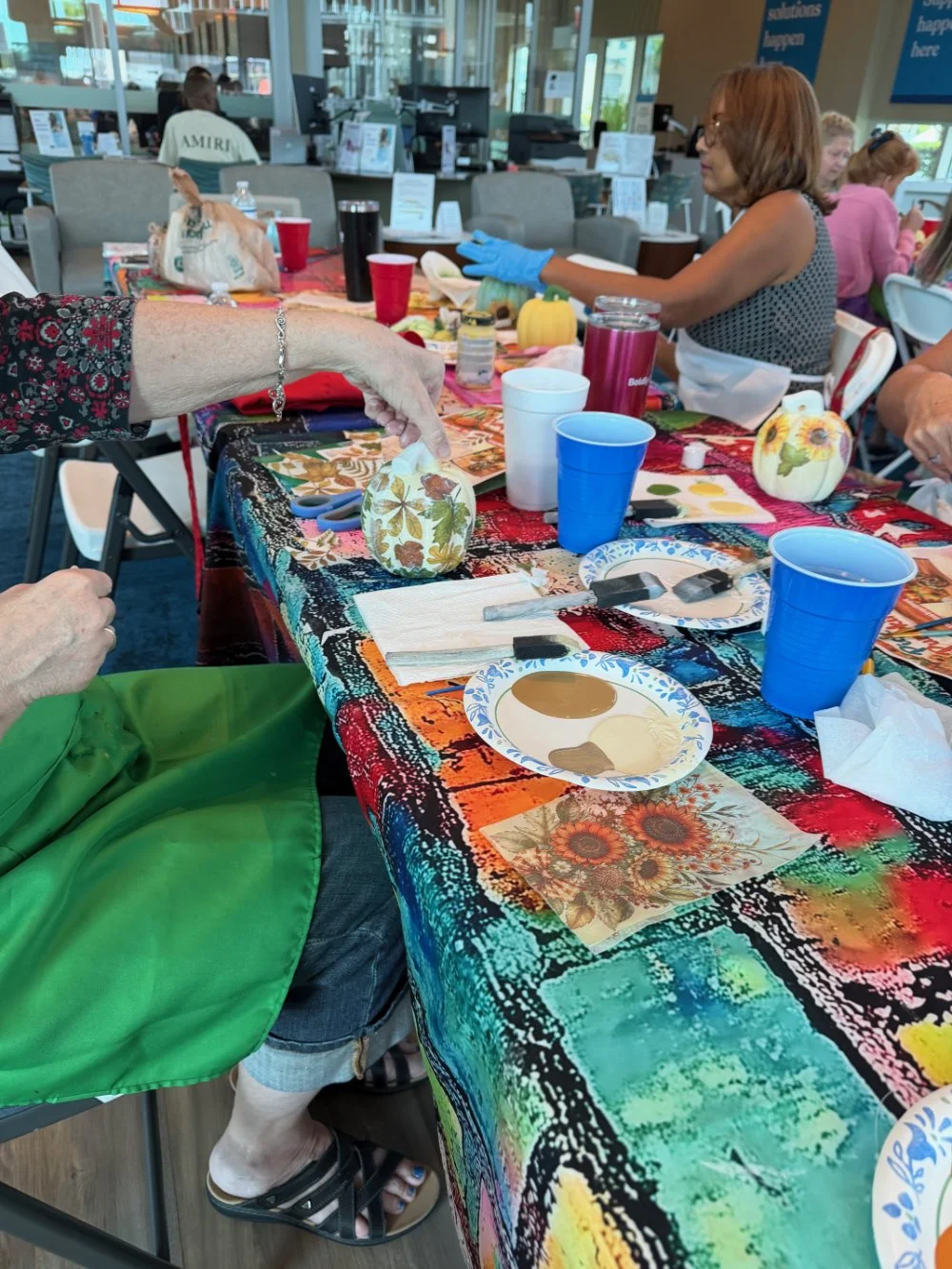 A colorful table decorated with autumn-themed items, including small pumpkins and fall motifs, set for a craft activity at a gathering in a bright, modern indoor space. Several people are engaged in decorating small pumpkin objects.