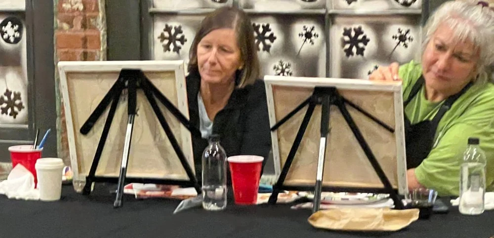 Two women painting on canvases set on easels during a holiday-themed event, with snowflake decorations in the background, and various supplies on the table.