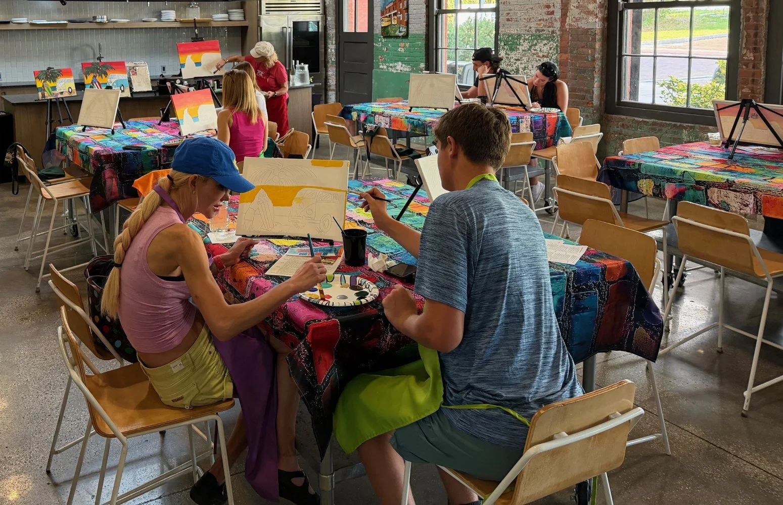 People painting canvases at a painting class in a decorated workshop with large windows, brick walls, and colorful tablecloths.