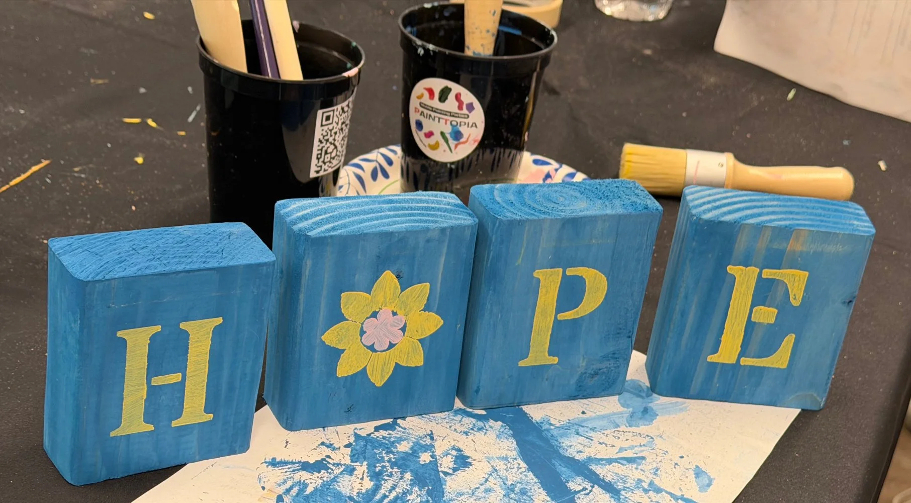 Blue painted wooden blocks spelling the word 'HOPE,' with the letter 'O' decorated with a yellow flower. Painting supplies and black cups with paintbrushes are in the background.
