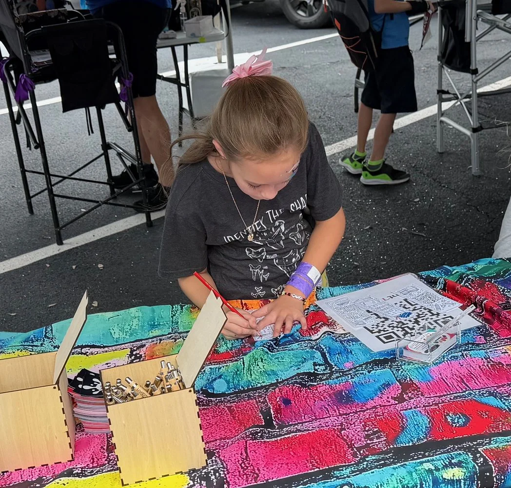 A young girl with a pink bow in her hair and a purple wristband is writing or drawing at a colorful table covered with a rainbow-colored cloth. There are QR codes and sign-up sheets on the table, along with two wooden containers holding small metal o