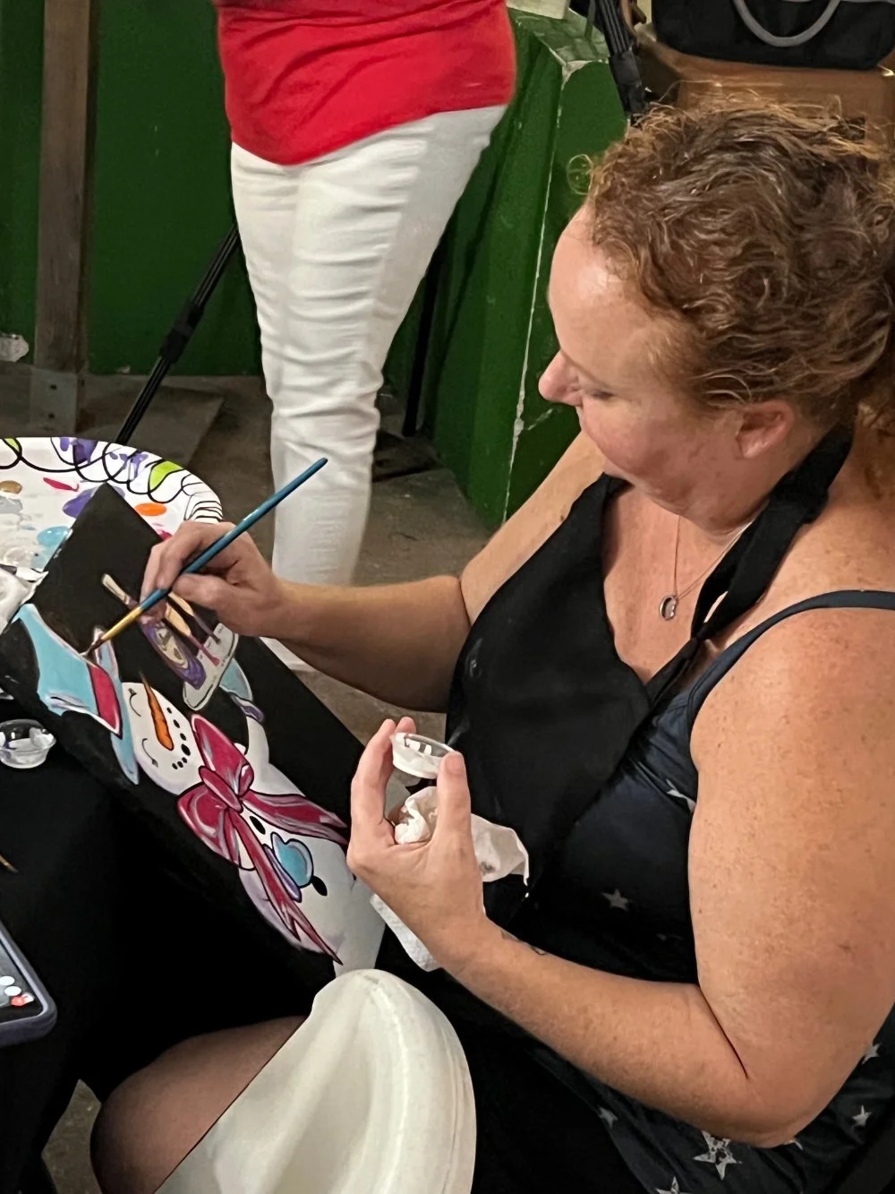 A woman painting a colorful snowman on a black surface, with painting supplies on the table.