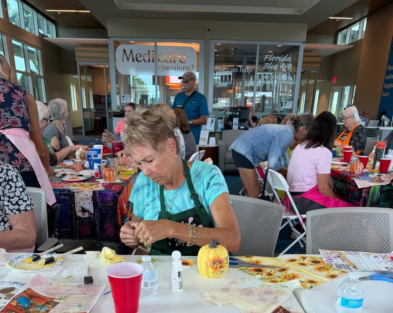 A group of people participating in a crafting event in a bright indoor space with large windows. They are seated at tables with colorful tablecloths, craft supplies, and decorations, including pumpkin-themed items. A sign in the background reads 'Med