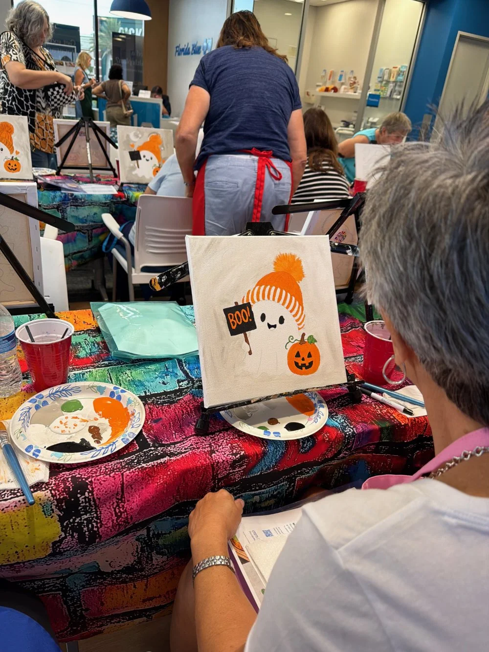A woman painting a Halloween-themed picture of a ghost with an orange hat, pumpkin, and a sign that says "BOO!" at a craft table during a group art event. The table has colorful tablecloth, paint palette, cups, and art supplies, and other people are 