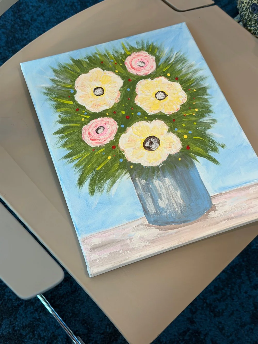 A painting of a flower bouquet in a blue vase with white, pink, and yellow flowers, placed on a light-colored surface.