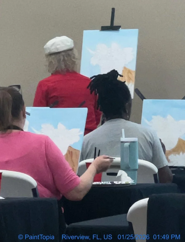 People seated at tables painting landscapes on canvases, with an instructor demonstrating at the front. The scene is indoors, and the participants are focused on their artwork.