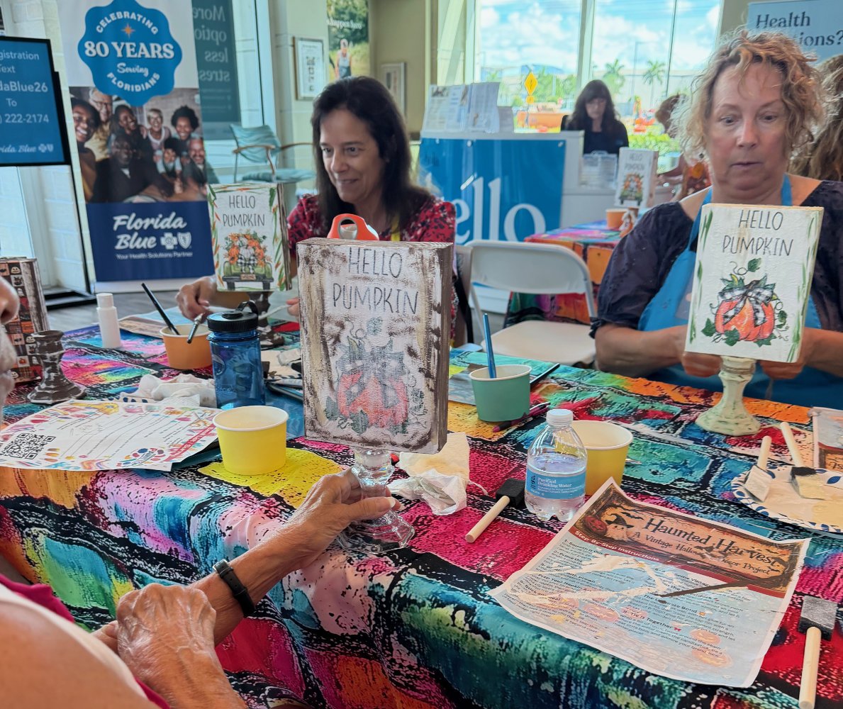 People sitting around a table decorated with a colorful tablecloth, crafting Halloween signs with pumpkins and the phrase 'Hello Pumpkin.' There are craft supplies on the table, including paint, brushes, paper, and a water bottle. In the background, 