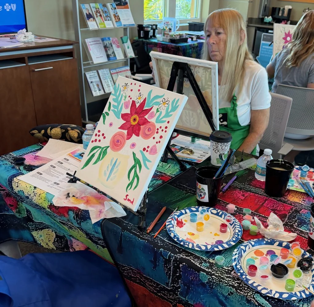 A woman with long blonde hair painting a floral artwork on a canvas at a colorful table with paints and brushes, surrounded by art supplies and a plastic water bottle.
