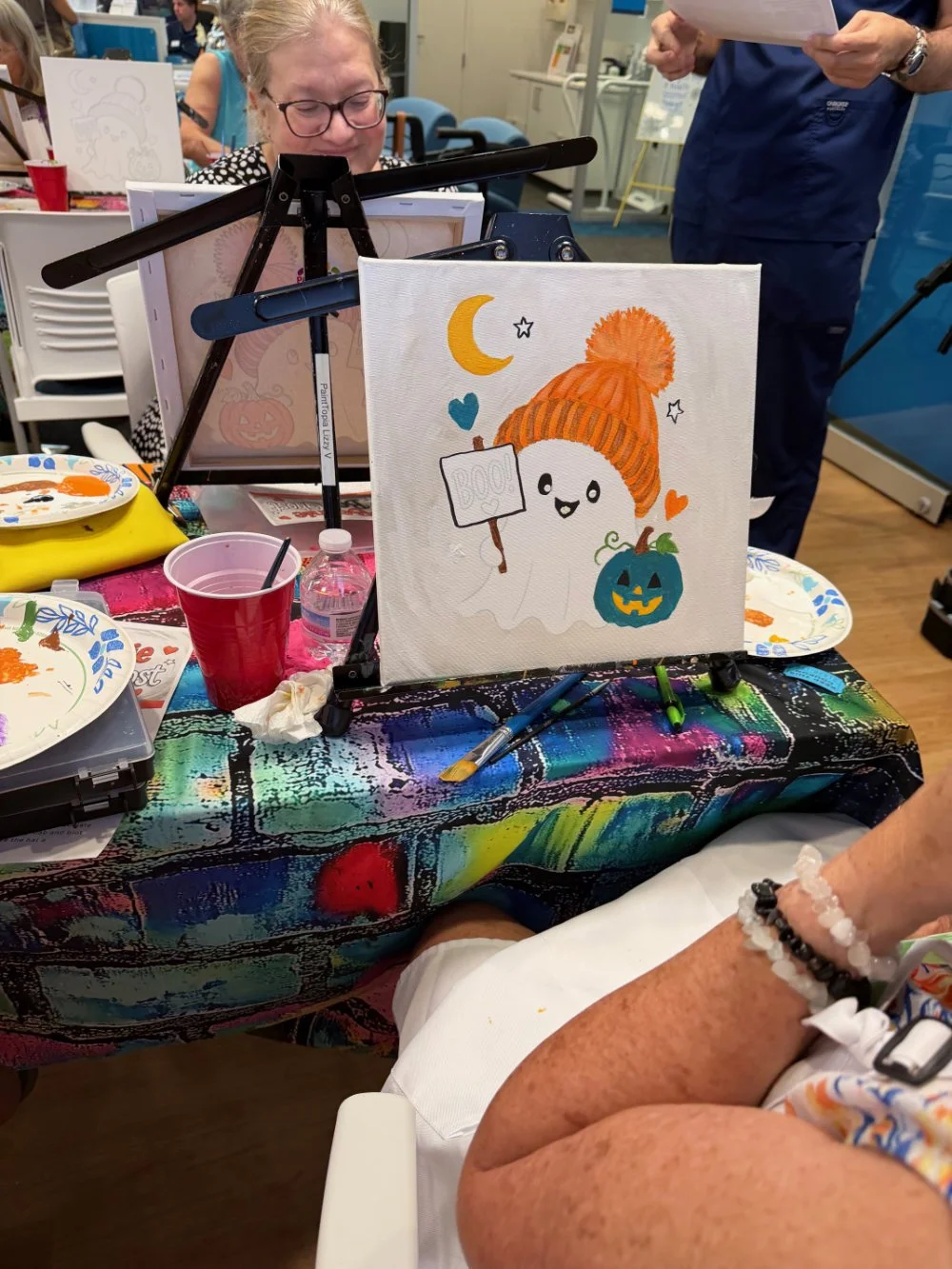 A small canvas with a cute Halloween ghost wearing an orange knit hat, holding a sign, surrounded by a moon, stars, hearts, and a pumpkin with a face, on a colorful table with painting supplies at an art event.