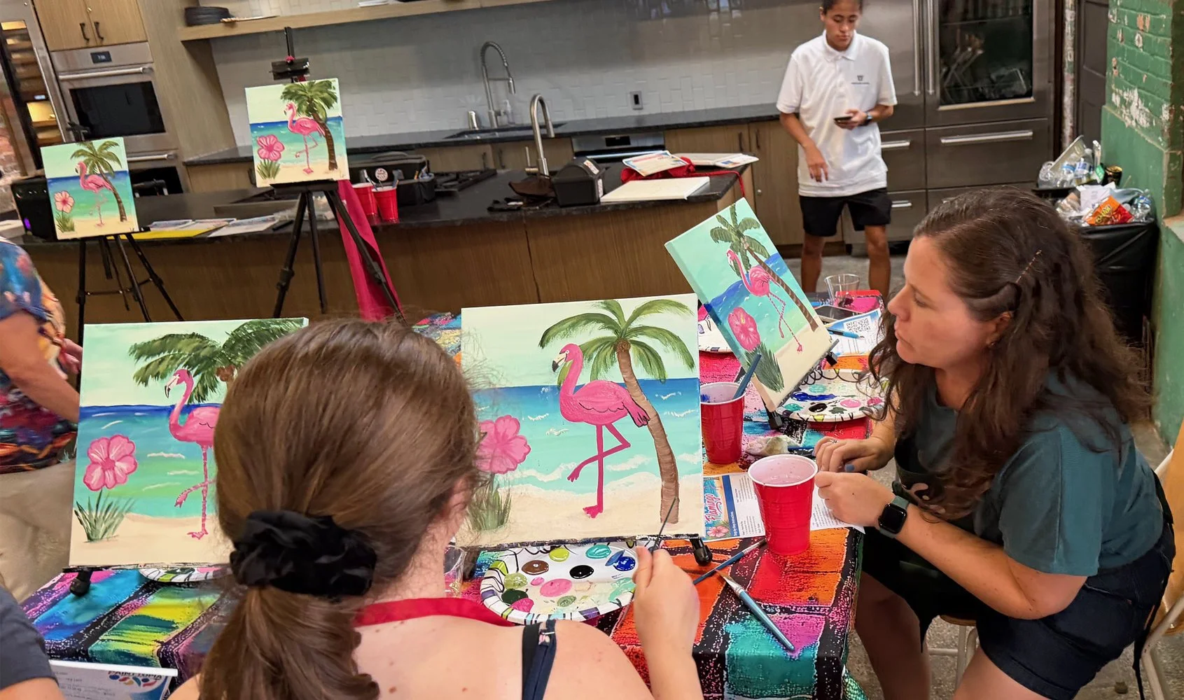 People painting tropical beach scenes with pink flamingos, palm trees, and pink flowers on small canvases at an art class.