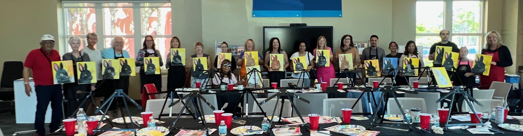 Group of people in a classroom holding up paintings of rabbits with a black background. The group is using painting easels and art supplies are on the tables.