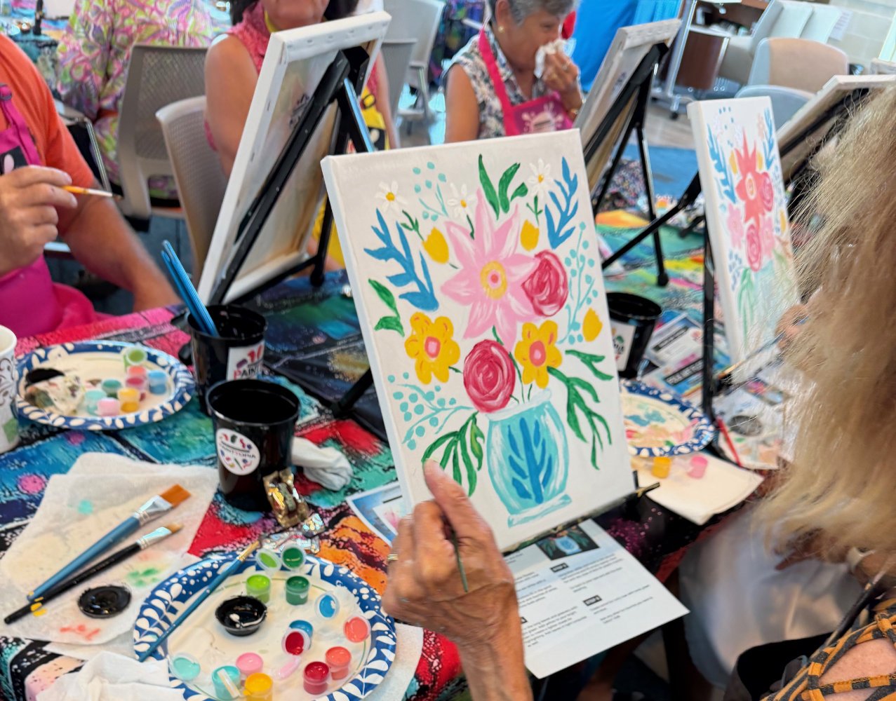 Women painting floral designs on canvases during a group art class, with paint palettes and brushes on the table.