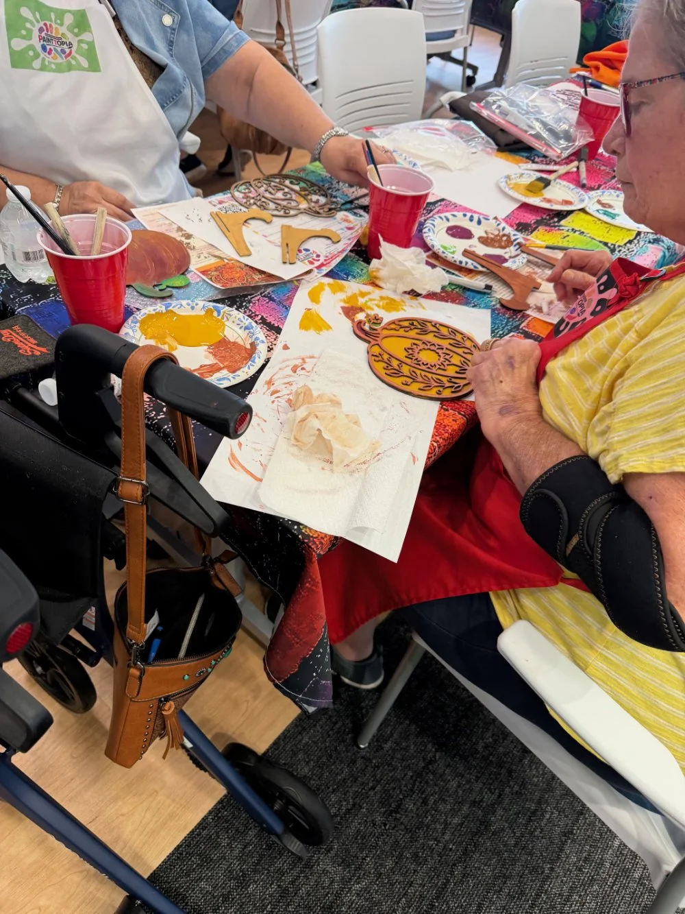 People participating in a paint decorating activity, working on orange and yellow pumpkin-shaped cutouts at a table covered with colorful paint supplies and artwork.