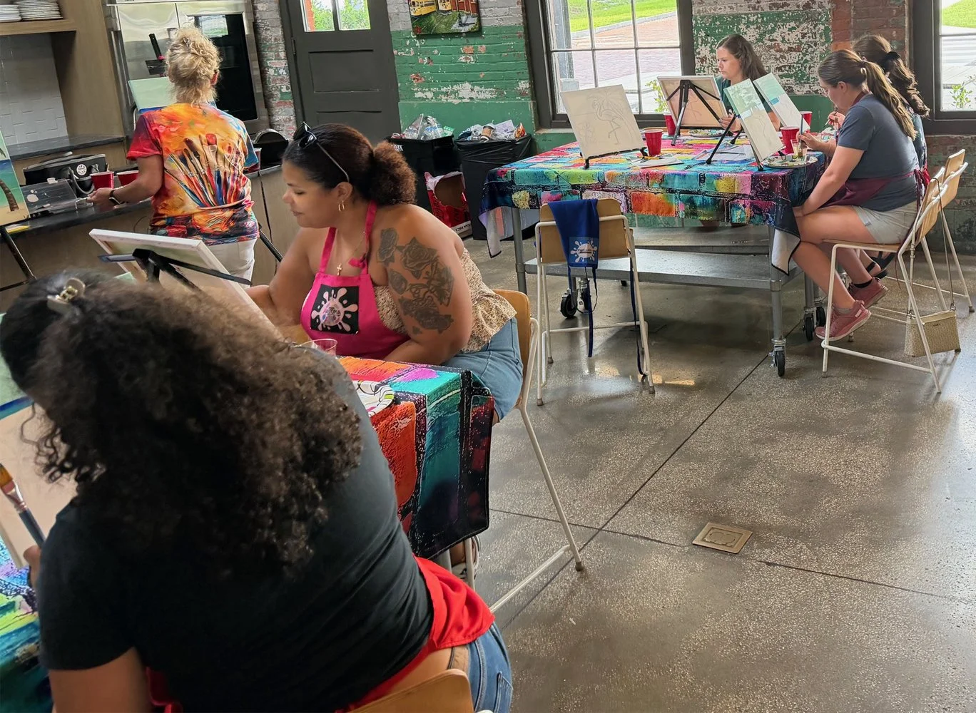 Women participating in a group painting class inside an art studio with paint supplies, easels, and colorful tables.