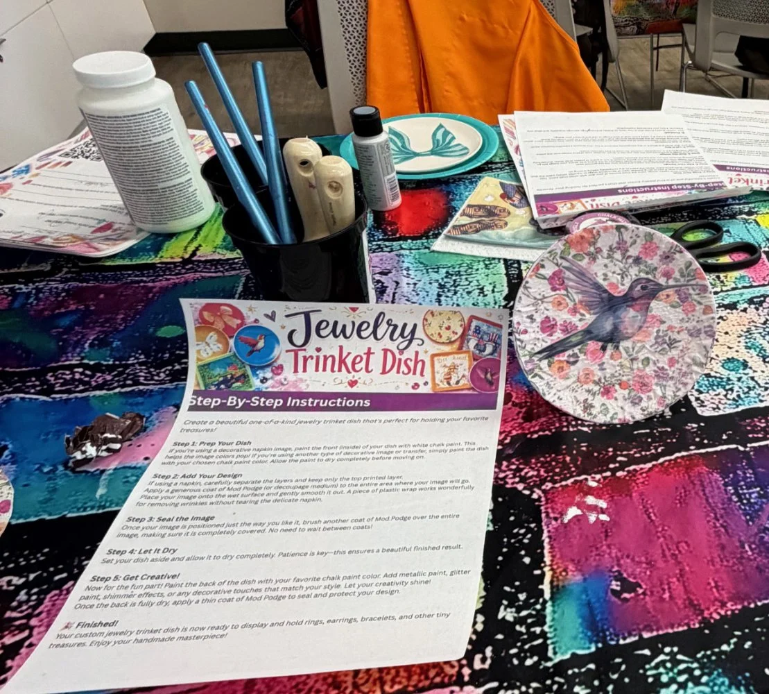 A colorful table covered with a jewelry trinket dish craft kit instructions, a black cup with paintbrushes, a spray paint bottle, pens, scissors, plates, and various papers.