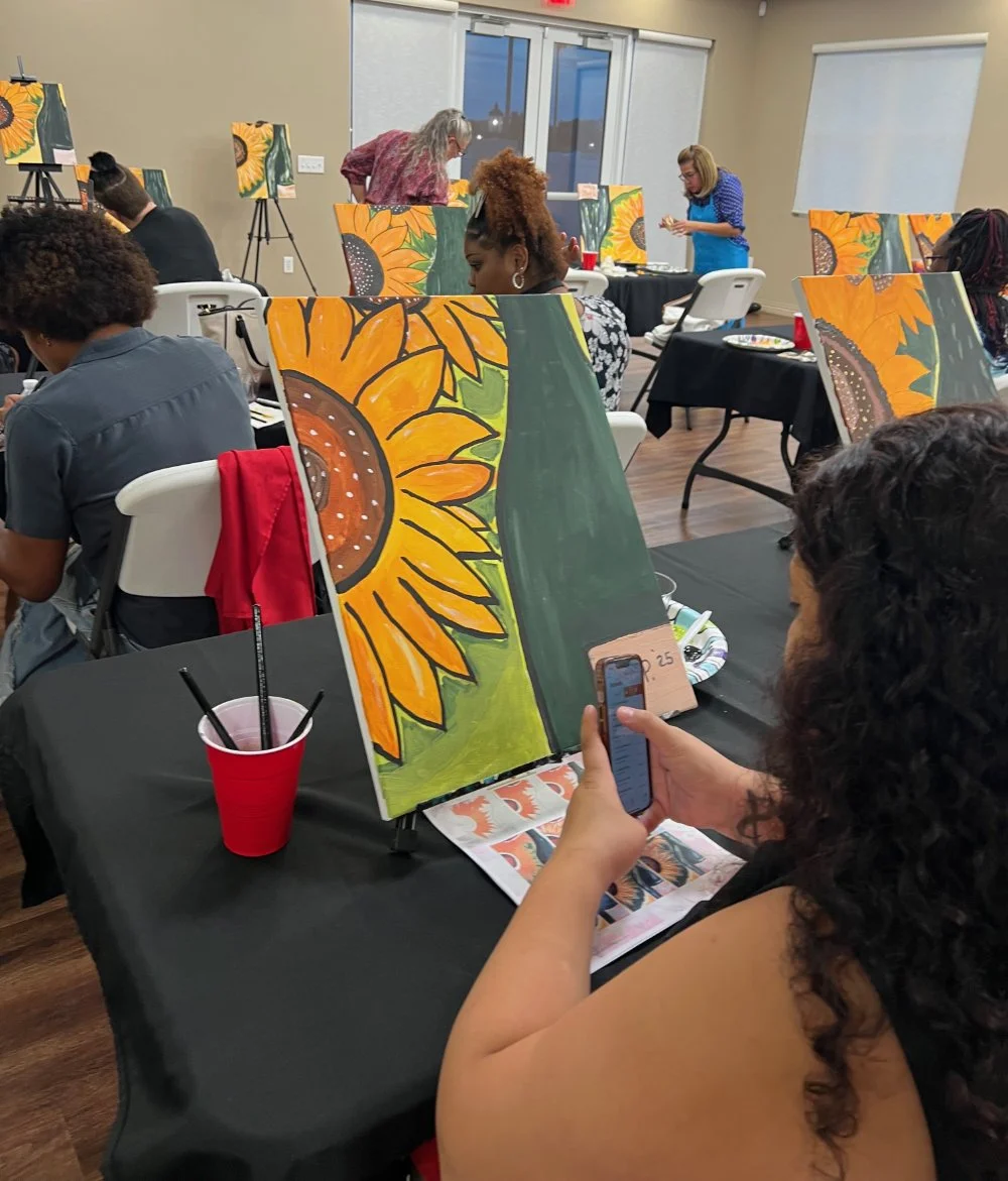 Participants in a painting class creating sunflower artwork; some are seated with canvases, while two instructors assist at the front of the room. A woman in the foreground is taking a photo of her sunflower painting with her phone.