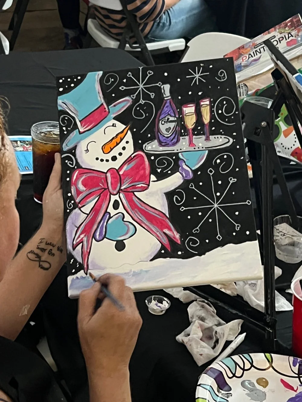 Painting of a festive snowman with a top hat, red bowtie, holding a tray of champagne glasses, surrounded by snowflakes and a black background.