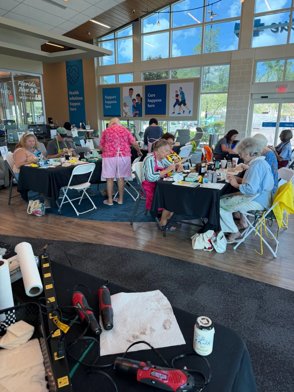 People sitting at tables in a bright, modern indoor space, possibly a community center or event area, with large windows and informational signs on the walls.