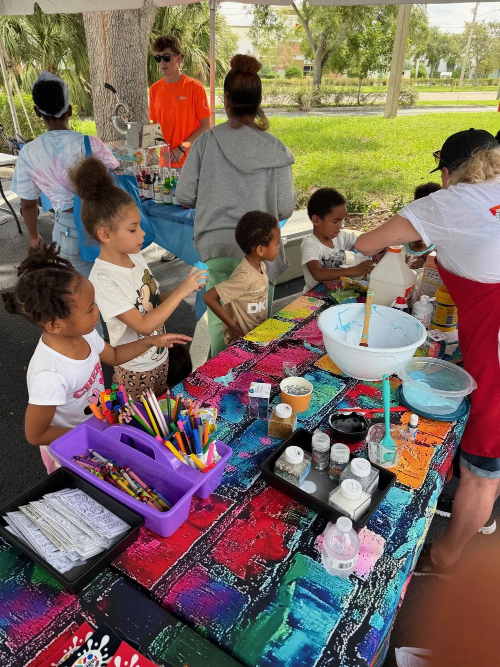 Children participating in a colorful outdoor arts and crafts activity under a canopy, with supplies such as markers, glitter, and paint on a vibrant table, guided by adults.