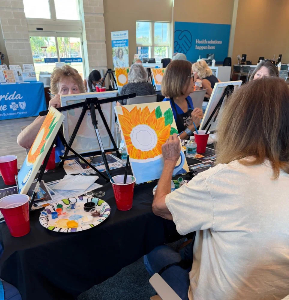 A group of people painting sunflower pictures at a painting event in a well-lit indoor space with large windows. There are red cups, painting supplies, and display boards with health-related messages in the background.