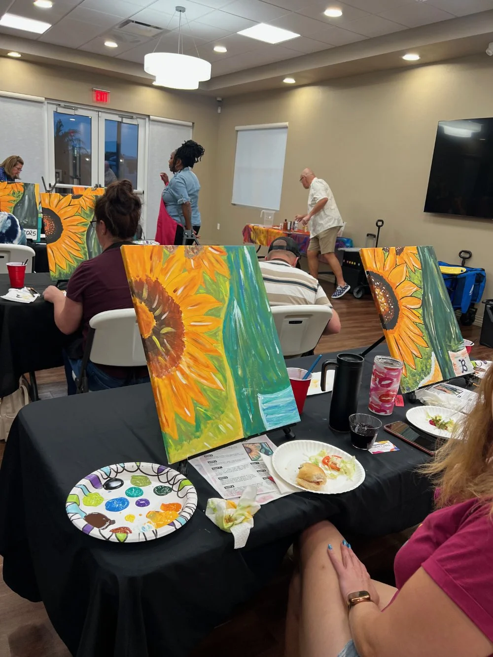 People participating in a sunflower painting event in a room with tables covered in black cloths, paintings of sunflowers on easels, and food on plates.