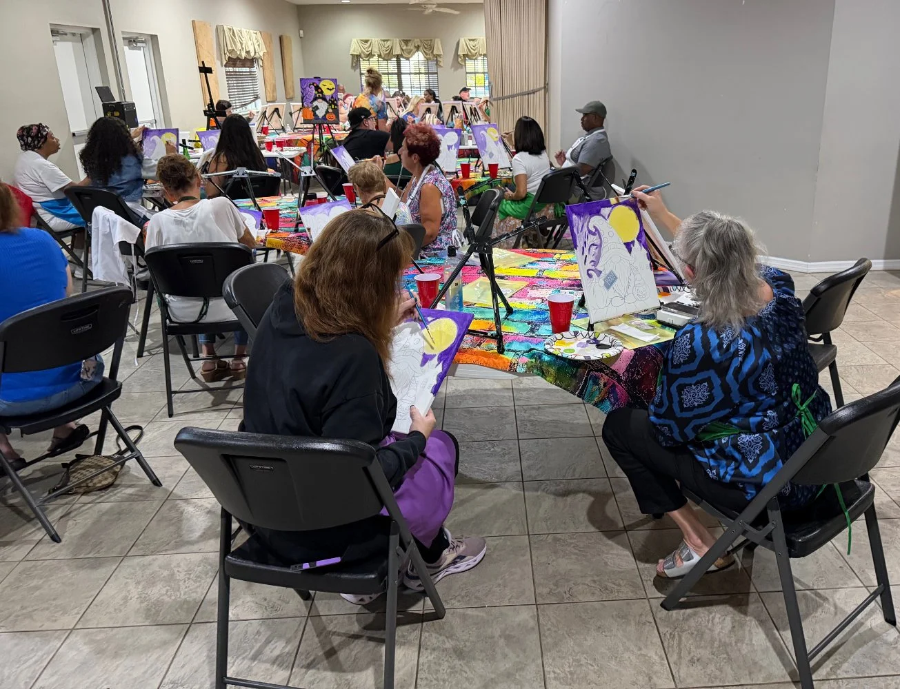 Group of people participating in a painting class, seated at a long, colorful table with painting supplies, focusing on their canvases with some paintings featuring a moon and a face.