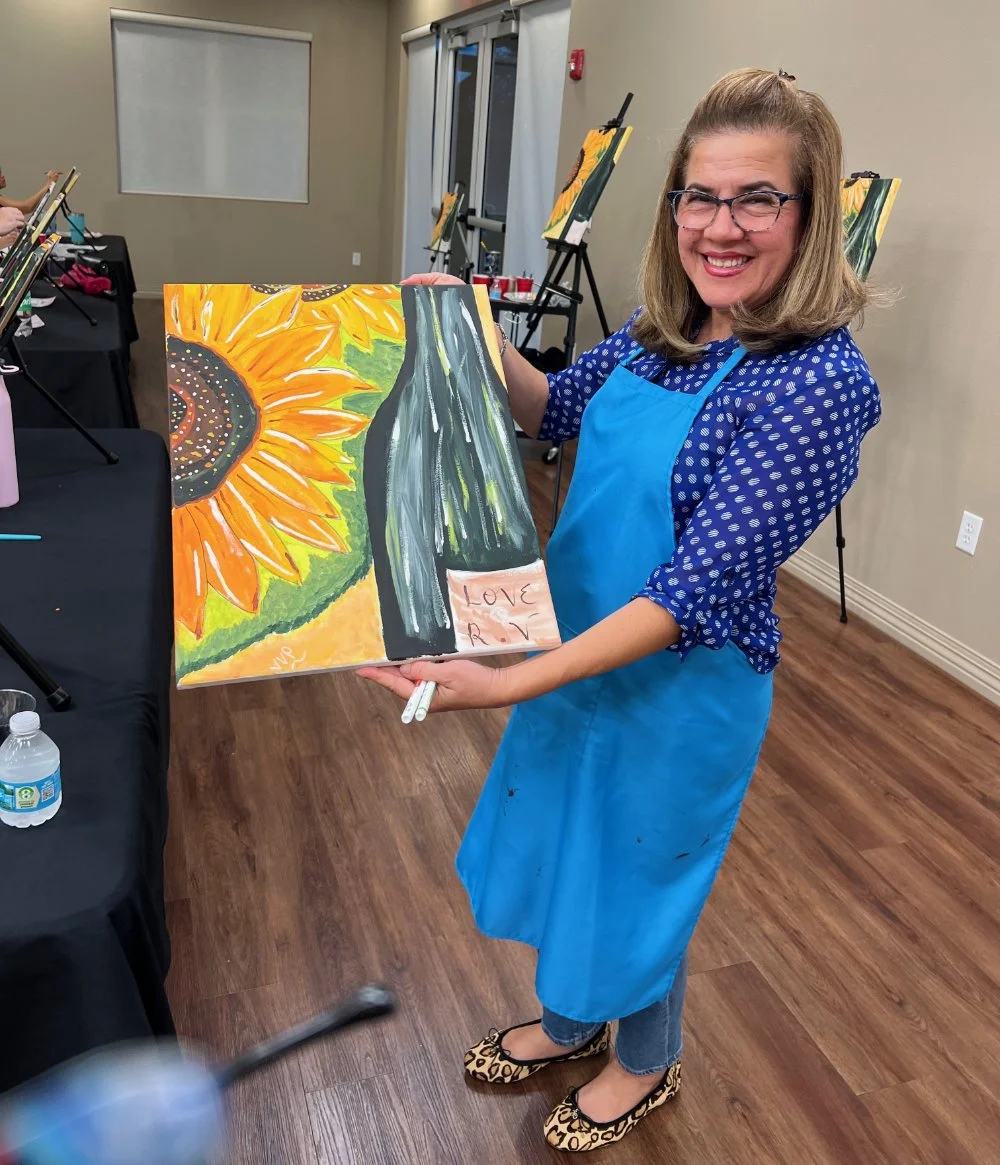 Woman in a blue apron holding a painting of a wine bottle with the words 'Love' and 'R' on it, standing in an art studio. The painting features a sunflower and vibrant colors.