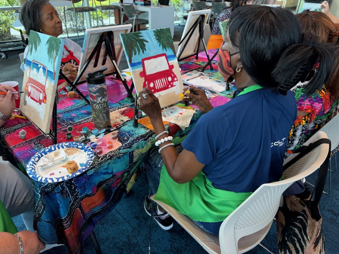 Group of women painting canvas panels with car and beach scene at a table covered with a colorful tablecloth, surrounded by art supplies and water bottles.