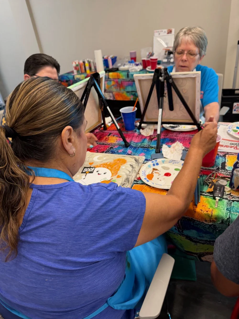 People engaged in a painting activity at a table covered with a colorful cloth, with painting supplies and art canvases.