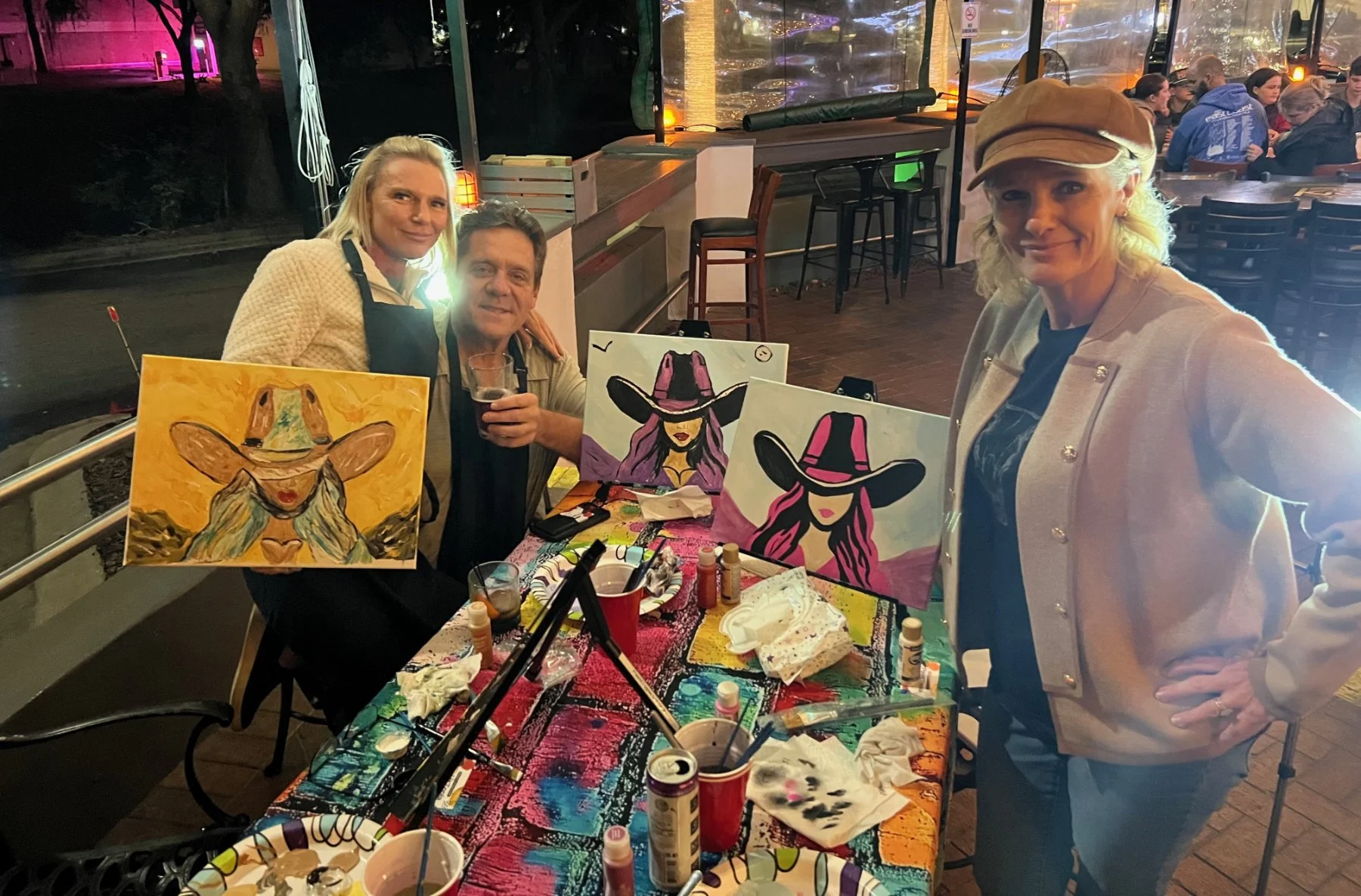 Group of three women at an art table creating and displaying painted portraits of women with large hats. One woman is seated, holding a drink, and two women are standing beside art displays. The table is covered with art supplies and partially completed paintings.