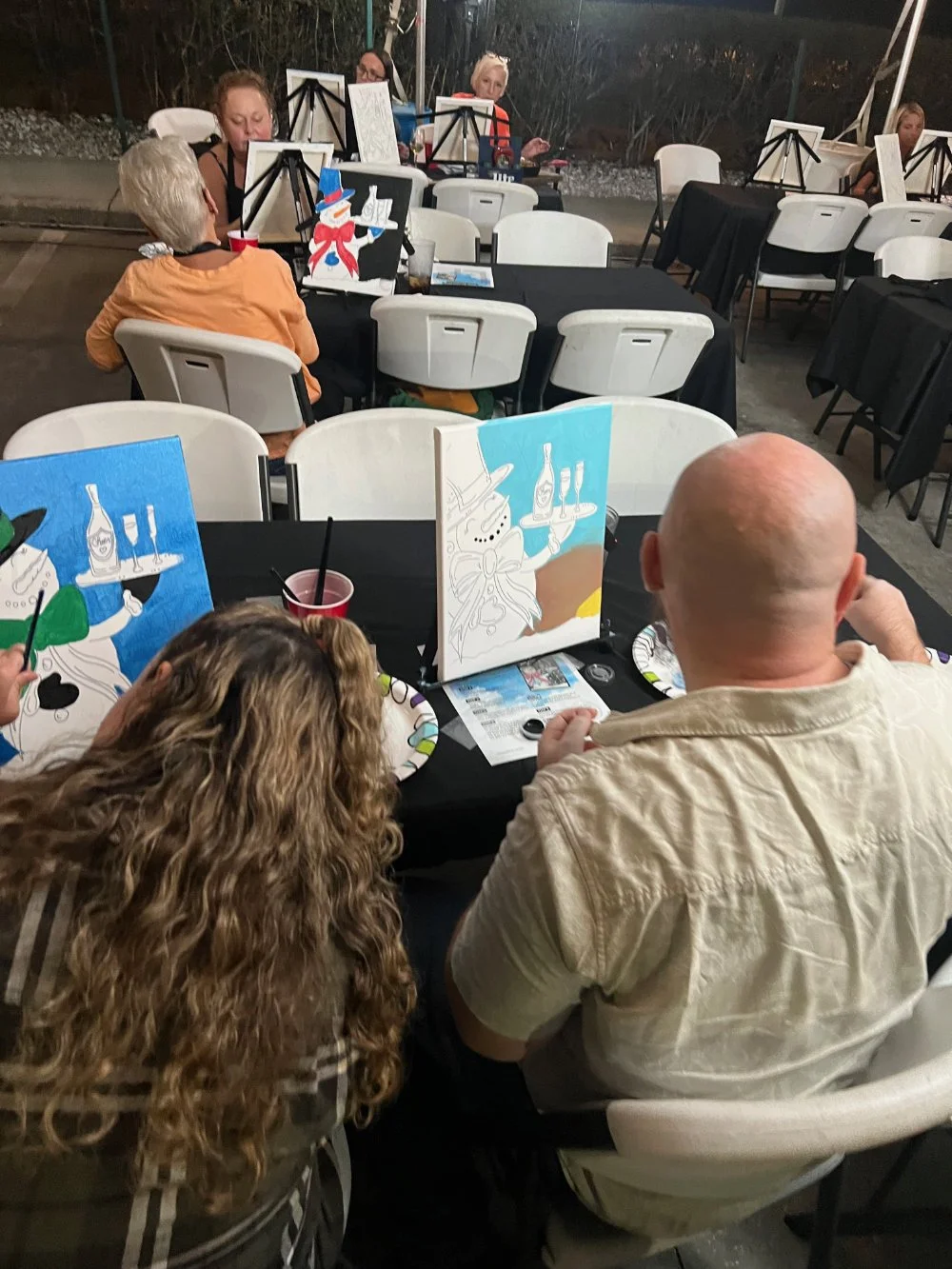 People painting at a social gathering, sitting around tables with painting projects of snowmen and snowflakes, with black tablecloths and disposable cups.