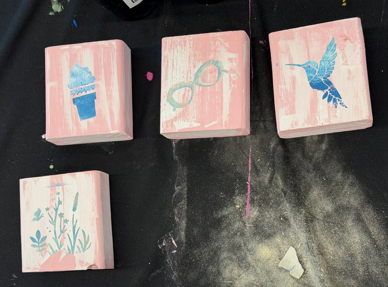 Four pink painted wooden blocks with blue stenciled images of an ice cream cone, glasses, a hummingbird, and plants arranged on a black surface.