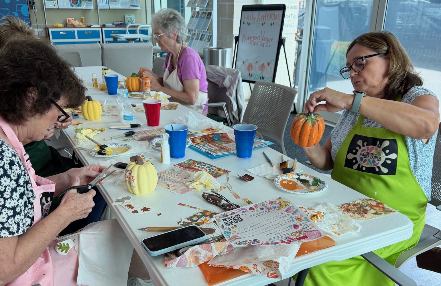 Women gathered at a table during a pumpkin painting craft activity, with craft supplies, painted pumpkins, and seasonal decorations.