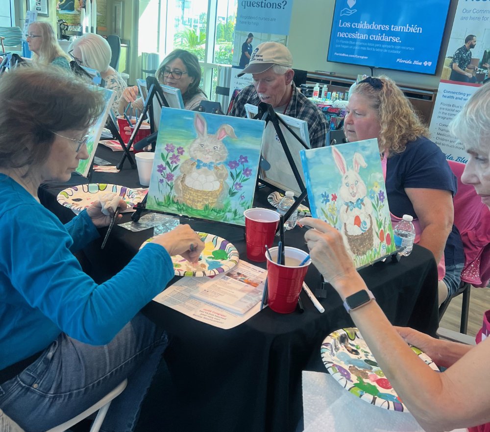 A group of people participating in a paint-and-sip class, painting pictures of rabbits with easels, plates, and cups on the table, with informational signs and a large screen in the background.