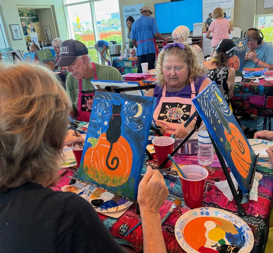 A group of people participating in a painting class at a recreational venue, painting Halloween-themed canvases featuring a black cat on a pumpkin with a moonlit night background.