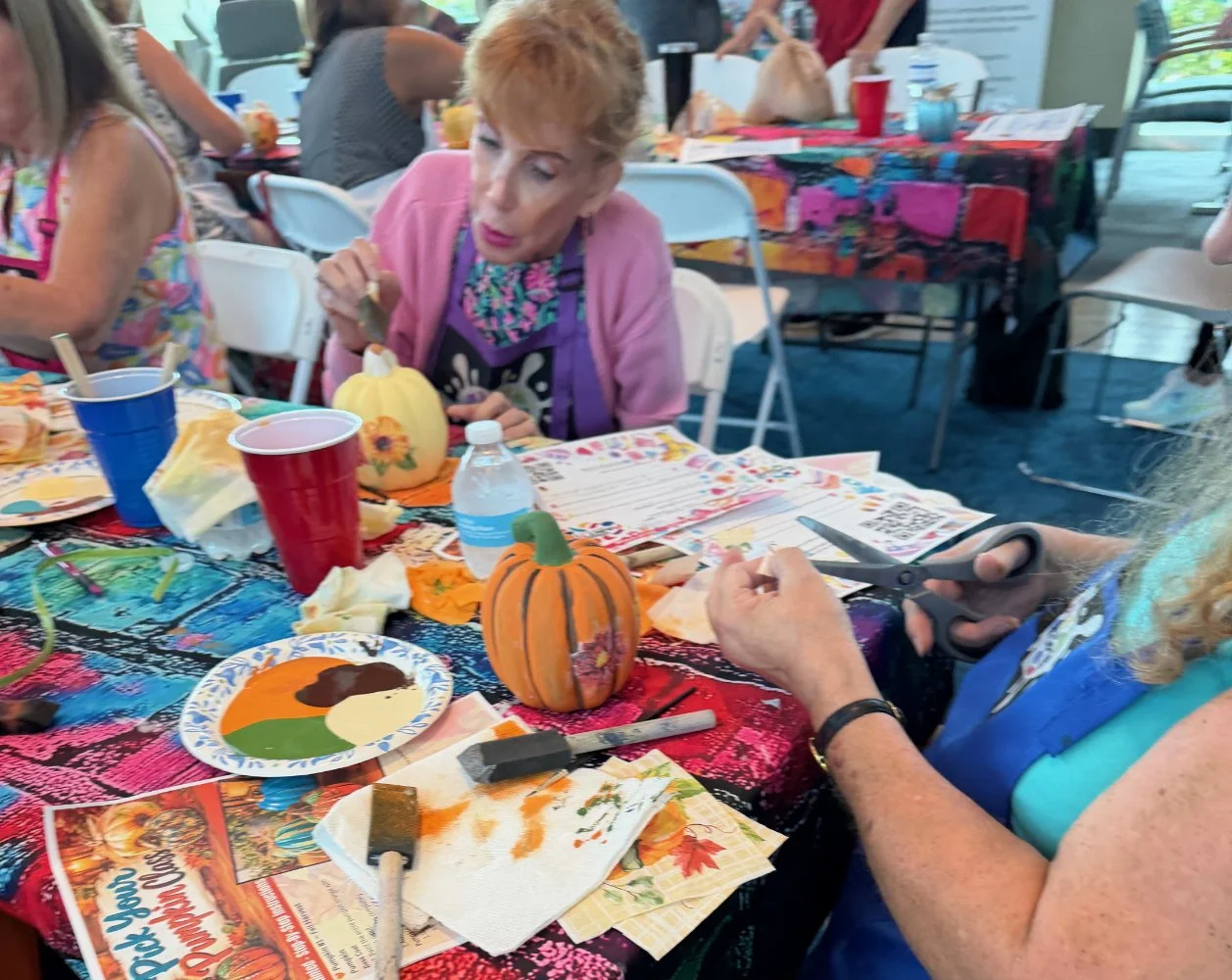 Women crafting and decorating small pumpkins at a table during a fall or Thanksgiving event, with craft supplies, water bottles, and colorful tablecloths.